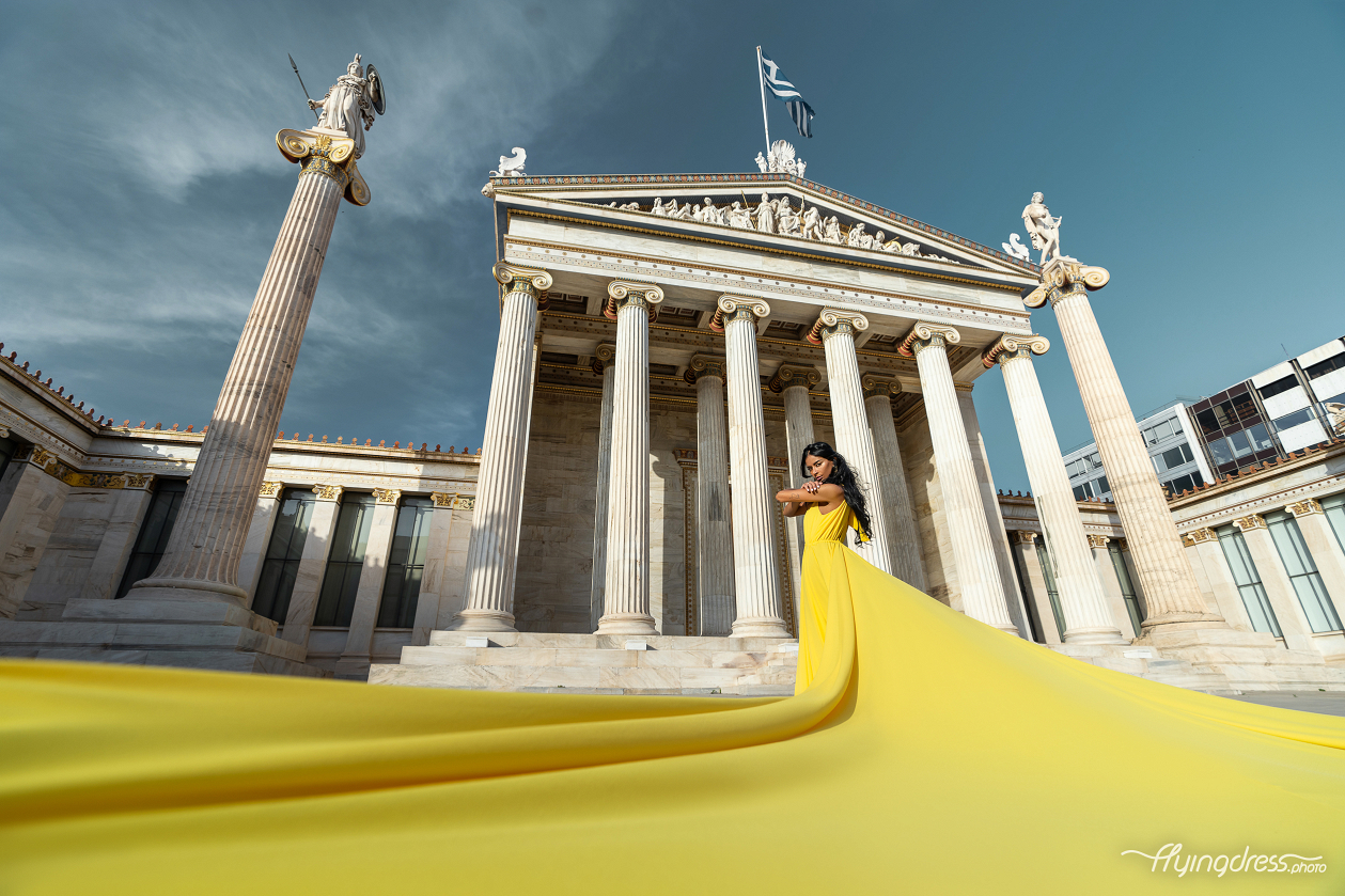 A woman in a flowing yellow dress poses in front of the grand columns of the Athens University, with the vibrant fabric of her dress trailing dramatically on the marble steps.